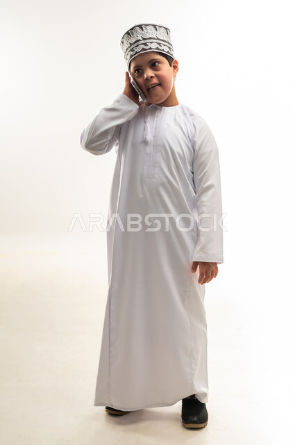 اجراء المكالمات الهاتفية، ايماءات السرور والسعادة، تعليم المعاقين على استخدام الأجهزة التقنية المتطورة، بورتريه لصبي عربي خليجي عماني مبتسم مصاب بمتلازمة داون يرتدي الدشداشة والكمة يحمل بيده الهاتف المحمول، صورة لكامل الجسم، خلفية بيضاء