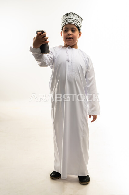 Browsing social media, developing the capabilities of people with disabilities, teaching the disabled to use advanced technical devices, portrait of a smiling Gulf Arab Omani boy with Down syndrome wearing a dishdasha and a cloak, holding a mobile phone in his hand, full body image, white background