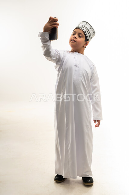 تعليم المعاقين على استخدام الأجهزة التقنية المتطورة، تنمية قدرات اصحاب الهمم، بورتريه لصبي عربي خليجي عماني مبتسم مصاب بمتلازمة داون يرتدي الدشداشة والكمة يحمل بيده الهاتف المحمول، تصفح مواقع التواصل الاجتماعي، صورة لكامل الجسم، خلفية بيضاء