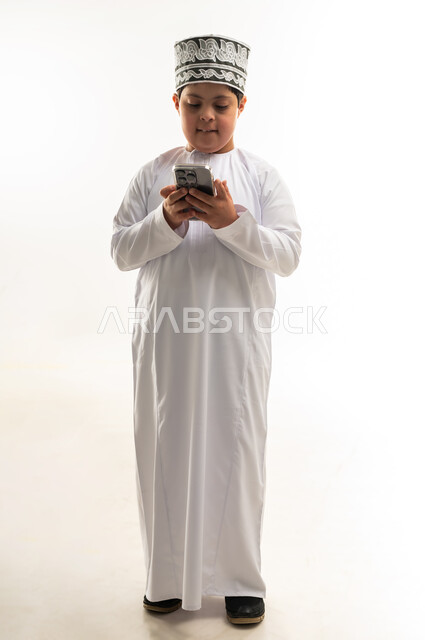 تنمية قدرات اصحاب الهمم، تعليم المعاقين على استخدام الأجهزة التقنية المتطورة، بورتريه لصبي عربي خليجي عماني مبتسم مصاب بمتلازمة داون يرتدي الدشداشة والكمة يحمل بيده الهاتف المحمول، تصفح مواقع التواصل الاجتماعي، صورة لكامل الجسم، خلفية بيضاء