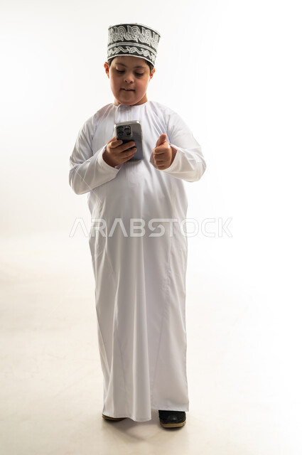 تصفح مواقع التواصل الاجتماعي، تعليم المعاقين على استخدام الأجهزة التقنية المتطورة، بورتريه لصبي عربي خليجي عماني مبتسم مصاب بمتلازمة داون يرتدي الدشداشة والكمة يحمل بيده الهاتف المحمول يرفع اصبع الابهام بايماءات القبول والموافقة، صورة لكامل الجسم، خلفية بيضاء