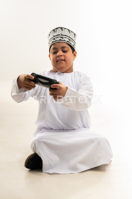 استخدام التقنيات الحديثة المسلية، قضاء أوقات الفراغ باللعب بألعاب الالكترونية الترفيهية، بورتريه لصبي عربي خليجي عمانيمصاب بمتلازمة داون يرتدي الدشاشة والكمة يحمل بيده عصا التحكم يرفع اصبع الابهام بايماءات الاعجاب والرضى، خلفية بيضاء