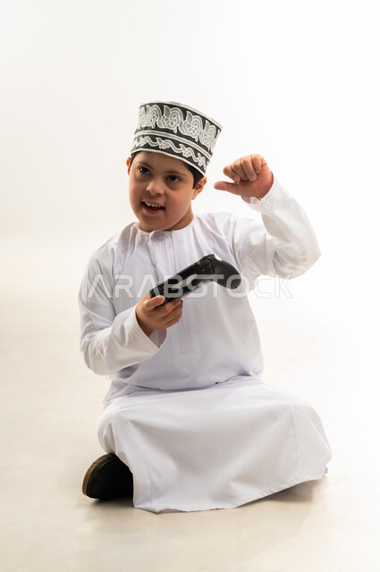 إيماءات السعادة والسرور، قضاء أوقات الفراغ باللعب بألعاب الالكترونية الترفيهية، استخدام التقنيات الحديثة المسلية، بورتريه لصبي عربي خليجي عمانيمصاب بمتلازمة داون يرتدي الدشاشة والكمة يحمل بيده عصا التحكم، خلفية بيضاء