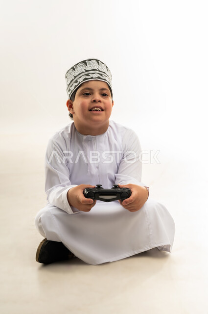 Spending free time playing entertaining electronic games, gestures of happiness and pleasure, using modern entertaining technologies, portrait of an Arab Gulf Omani boy with Down syndrome wearing a screen and a mask, holding a joystick in his hand, white background