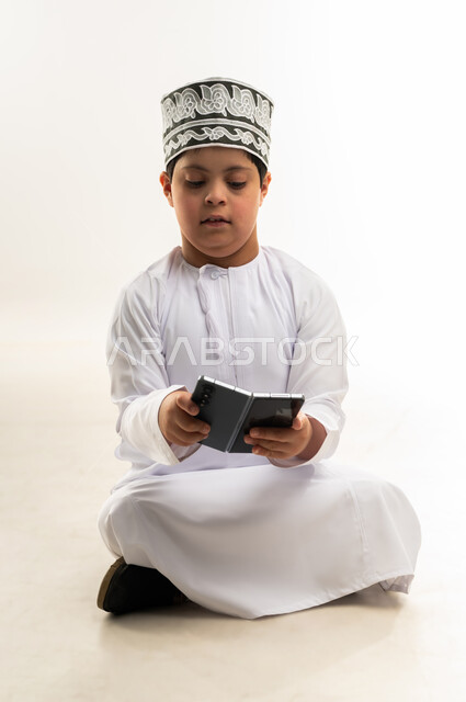 Teaching the disabled to use advanced technical devices, developing the capabilities of people with disabilities, portrait of a smiling Gulf Arab Omani boy with Down syndrome wearing a dishdasha and a cloak, holding a mobile phone in his hand, browsing social media, white background