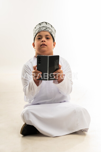 تنمية قدرات اصحاب الهمم، تعليم المعاقين على استخدام الأجهزة التقنية المتطورة، بورتريه لصبي عربي خليجي عماني مبتسم مصاب بمتلازمة داون يرتدي الدشداشة والكمة يحمل بيده الهاتف المحمول، تصفح مواقع التواصل الاجتماعي، خلفية بيضاء