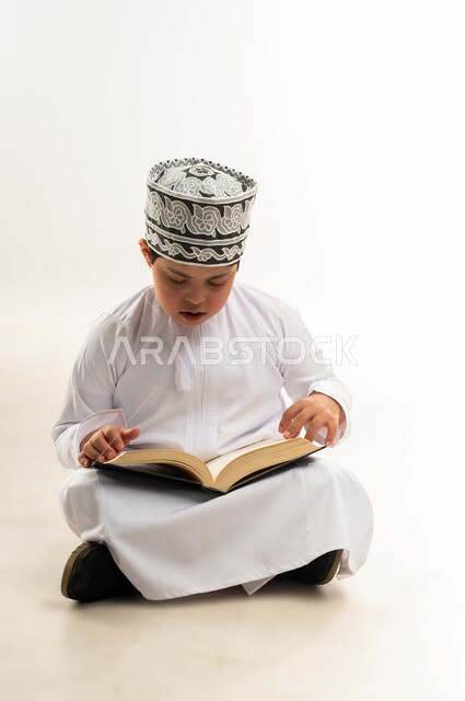 مفهوم الاجتهاد في العبادة، تلاوة المصحف الشريف، قراءة كتاب الله وحفظه وتجويده، ايماءات التركيز والاندماج، اسلوب حياة ذوي الهمم، بورتريه لصبي عربي خليجي عماني يرتدي الدشداشة والكمة يحمل بيده القرآن، خلفية بيضاء