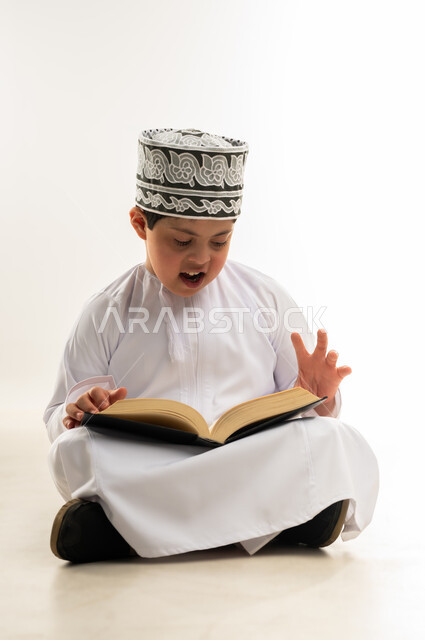 تلاوة المصحف الشريف، قراءة كتاب الله وحفظه وتجويده، ايماءات التركيز والاندماج، مفهوم الاجتهاد في العبادة، اسلوب حياة ذوي الهمم، بورتريه لصبي عربي خليجي عماني يرتدي الدشداشة والكمة يحمل بيده القرآن، خلفية بيضاء