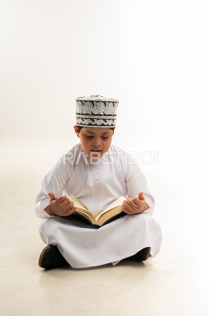 Gestures of concentration and integration, reading the Book of Allah, memorizing it and reciting it with Tajweed, reciting the Holy Quran, the concept of diligence in worship, the lifestyle of people with disabilities, a portrait of an Arab Gulf Omani boy wearing a dishdasha and a kummah holding the Quran in his hand, white background