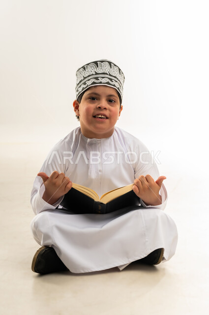 قراءة كتاب الله وحفظه وتجويده، مفهوم الاجتهاد في العبادة، تلاوة المصحف الشريف، ايماءات التركيز والاندماج، اسلوب حياة ذوي الهمم، بورتريه لصبي عربي خليجي عماني يرتدي الدشداشة والكمة يحمل بيده القرآن، خلفية بيضاء