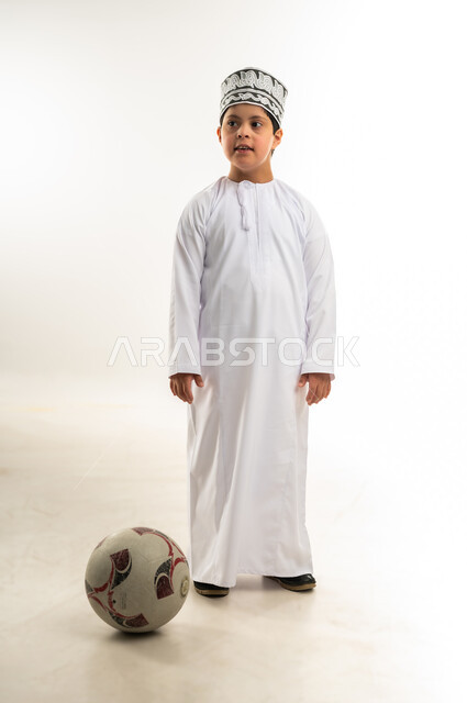 Developing physical skills for people with disabilities, lifestyle of people with disabilities, practicing favorite hobbies, portrait of an Arab Gulf Omani boy with Down syndrome wearing a dishdasha and a kummah playing football, full body image, white background