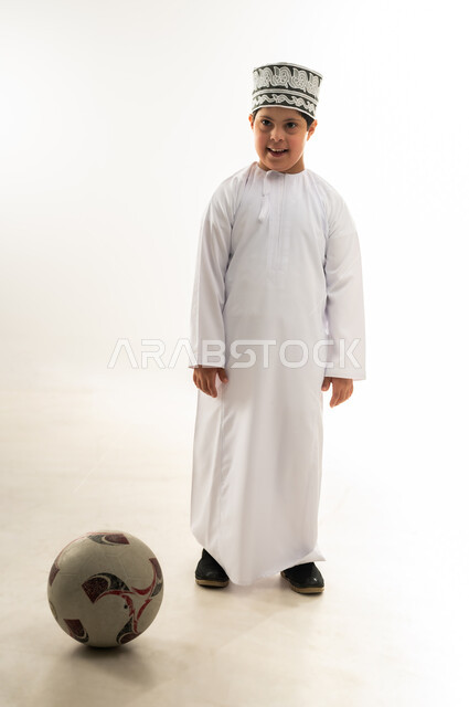 Practicing favorite hobbies, developing and improving physical skills for people with disabilities, lifestyle of people with disabilities, portrait of an Arab Gulf Omani boy with Down syndrome wearing a dishdasha and a kimono playing football, full body image, white background