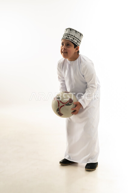 تنمية وتطوير المهارات البدنية لذوي الإعاقة، ممارسة الهوايات المفضلة، اسلوب حياة ذوي الهمم، بورتريه لصبي عربي خليجي عماني مصاب بمتلازمة داون يرتدي الدشداشة والكمة يحمل بيده كرة القدم، صورة لكامل الجسم، خلفية بيضاء