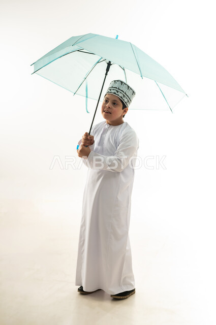 الحماية من الأمطار في الشتاء، التظلل من أشعة الشمس، بورتريه لصبي عربي خليجي عماني مبتسم مصاب بمتلازمة داون يرتدي الدشداشة والكمة يحمل بيده مظلة زرقاء، دمج ذوي الهمم بالمجتمع العماني، صورة لكامل الجسم، خلفية بيضاء