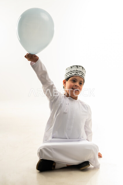 مفهوم الترفيه عن النفس والتسلية، اسلوب حياة ذوي الهمم، بورتريه لولد عربي خليجي عماني مصاب بمتلازمة داون يرتدي الدشداشة والكمة يحمل بالون باللون الازرق يرفع يدهبايماءات السرور والسعادة، دعم المجتمع العماني لذوي الاحتياجات الخاصة، خلفية بيضاء