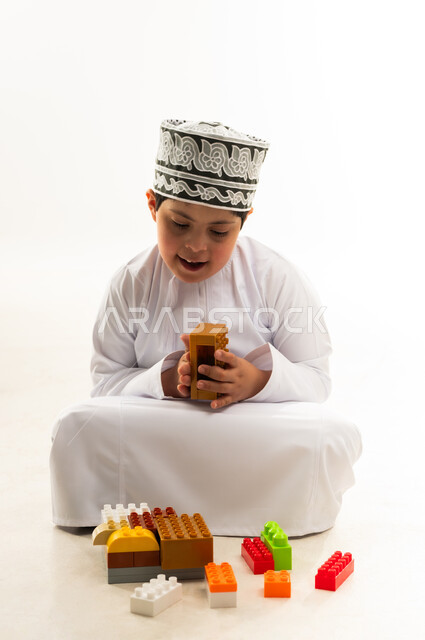 اهتمام المجتمع العماني بأصحاب الهمم, نشاطات ترفيهية لذوي الاحتياجات الخاصة, بورتريه لصبي عربي خليجي عماني مصاب بمتلازمة داون يرتدي الدشداشة والكمة يقوم بتركيب قطع المكعبات، تنمية القدرات الذهنية للمعوقين، خلفية بيضاء