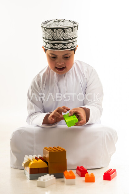 تنمية القدرات الذهنية للمعوقين، اهتمام المجتمع العماني بأصحاب الهمم, نشاطات ترفيهية لذوي الاحتياجات الخاصة, بورتريه لصبي عربي خليجي عماني مصاب بمتلازمة داون يرتدي الدشداشة والكمة يقوم بتركيب قطع المكعبات، خلفية بيضاء