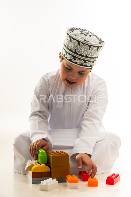 نشاطات ترفيهية لذوي الاحتياجات الخاصة, اهتمام المجتمع العماني بأصحاب الهمم, بورتريه لصبي عربي خليجي عماني مصاب بمتلازمة داون يرتدي الدشداشة والكمة يقوم بتركيب قطع المكعبات، تنمية القدرات الذهنية للمعوقين، خلفية بيضاء