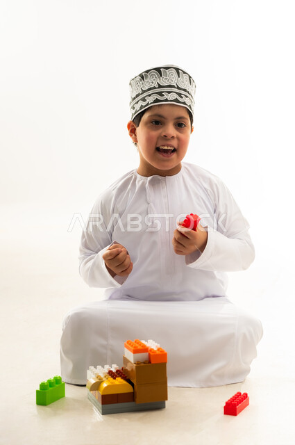 ايماءات السرور والسعادة، تنمية القدرات الذهنية للمعوقين، اهتمام المجتمع العماني بأصحاب الهمم, نشاطات ترفيهية لذوي الاحتياجات الخاصة, بورتريه لصبي عربي خليجي عماني مصاب بمتلازمة داون يرتدي الدشداشة والكمة يقوم بتركيب قطع المكعبات، خلفية بيضاء