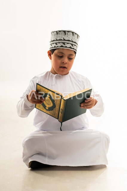 مفهوم الاجتهاد في العبادة، تلاوة المصحف الشريف، قراءة كتاب الله وحفظه وتجويده، ايماءات التركيز والاندماج، اسلوب حياة ذوي الهمم، بورتريه لصبي عربي خليجي عماني يرتدي الدشداشة والكمة يحمل بيده القرآن، خلفية بيضاء