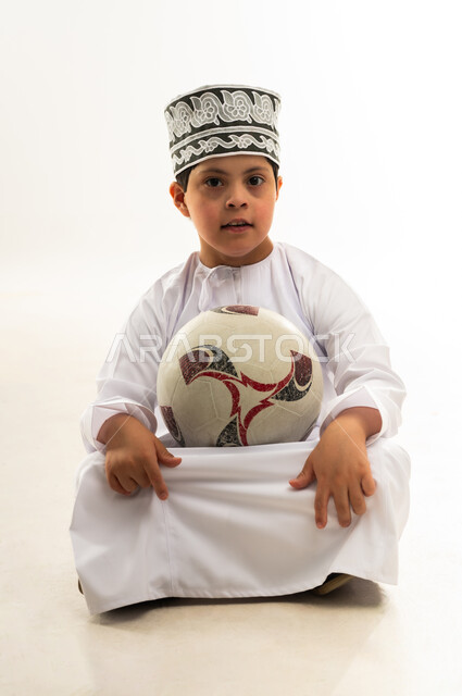 النظر إلى الكاميرا بتعابير السرور والسعادة، تنمية الهوايات الرياضية لذوي الإعاقة، الشغف في متابعه المباريات، اسلوب حياة ذوي الهمم، بورتريه لصبي عربي خليجي عماني مصاب بمتلازمة داون يرتدي الدشداشة والكمة يحمل كرة القدم، خلفية بيضاء