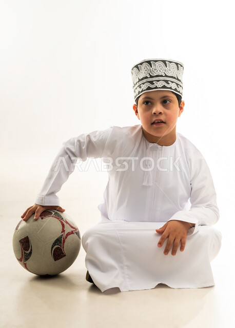 الشغف في متابعه المباريات، اسلوب حياة ذوي الهمم، تنمية الهوايات الرياضية لذوي الإعاقة, بورتريه لصبي عربي خليجي عماني مصاب بمتلازمة داون يرتدي الدشداشة والكمة يجلس بجانب كرة القدم، صورة لكامل الجسم، خلفية بيضاء