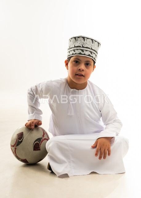 اسلوب حياة ذوي الهمم، النظر إلى الكاميرا بتعابير السرور والسعادة، الشغف في متابعه المباريات، تنمية الهوايات الرياضية لذوي الإعاقة, بورتريه لصبي عربي خليجي عماني مصاب بمتلازمة داون يرتدي الدشداشة والكمة يجلس بجانب كرة القدم، خلفية بيضاء