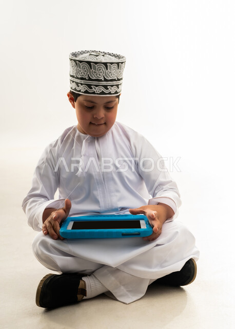 اسلوب حياة أصحاب الهمم، استخدام التكنولوجيا للتسلية، الترفيه على الأجهزة المحمولة، لعب ألعاب الفيديو الممتعة، بورتريه لصبي عربي خليجي عماني مبتسم مصاب بمتلازمة داون يرتدي الدشداشة والكمة يحمل بيده التابلت، خلفية بيضاء