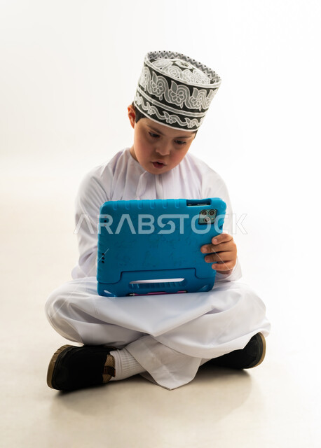 الترفيه على الأجهزة المحمولة، اسلوب حياة أصحاب الهمم، استخدام التكنولوجيا للتسلية، لعب ألعاب الفيديو الممتعة، بورتريه لصبي عربي خليجي عماني مبتسم مصاب بمتلازمة داون يرتدي الدشداشة والكمة يحمل بيده التابلت، خلفية بيضاء