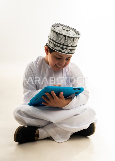 استخدام التكنولوجيا للتسلية، اسلوب حياة أصحاب الهمم، الترفيه على الأجهزة المحمولة، لعب ألعاب الفيديو الممتعة، بورتريه لصبي عربي خليجي عماني مبتسم مصاب بمتلازمة داون يرتدي الدشداشة والكمة يحمل بيده التابلت، خلفية بيضاء