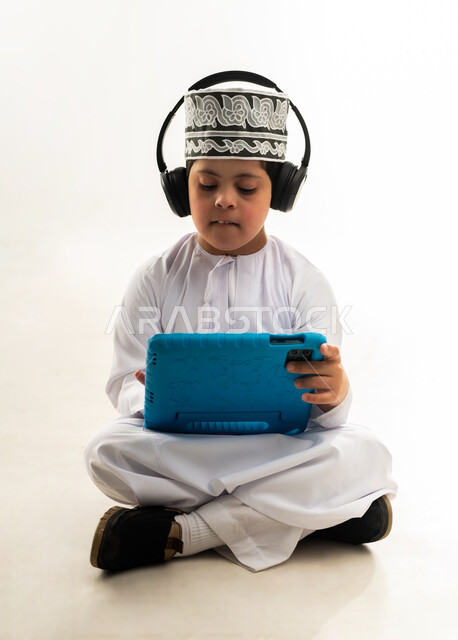 الترفيه على الأجهزة المحمولة، اسلوب حياة أصحاب الهمم، استخدام التكنولوجيا للتسلية، لعب ألعاب الفيديو الممتعة، بورتريه لصبي عربي خليجي عماني مبتسم مصاب بمتلازمة داون يرتدي الدشداشة والكمة يضع السماعات يحمل بيده التابلت، خلفية بيضاء