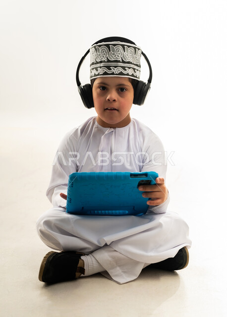 لعب ألعاب الفيديو الممتعة، الترفيه على الأجهزة المحمولة، اسلوب حياة أصحاب الهمم، استخدام التكنولوجيا للتسلية، بورتريه لصبي عربي خليجي عماني مبتسم مصاب بمتلازمة داون يرتدي الدشداشة والكمة يضع السماعات يحمل بيده التابلت، خلفية بيضاء