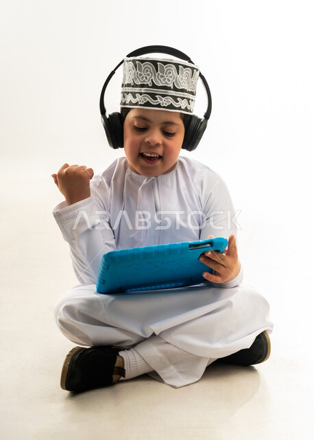 لعب ألعاب الفيديو الممتعة، الترفيه على الأجهزة المحمولة، استخدام التكنولوجيا للتسلية، بورتريه لصبي عربي خليجي عماني مبتسم مصاب بمتلازمة داون يرتدي الدشداشة والكمة يضع السماعات يحمل التابلت يرفع يده بايماءات الحماس، خلفية بيضاء