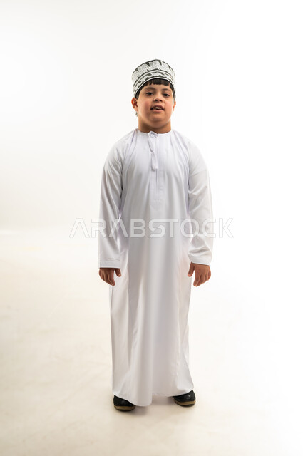 الوقوف باستقامة وانضباط، مفهوم الاناقة و الاهتمام بالمظهر الخارجي, بورتريه لولد عربي خليجي عماني يرتدي الدشداشة و الكمه ينظر إلى الكاميرا بإيماءات السرور والسعادة، صورة الجسم بالطول الكامل, خلفية بيضاء