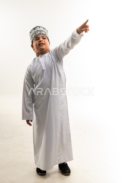 تحديد موقع معين، مفهوم الاناقة والاهتمام بالمظهر الخارجي, بورتريه لولد عربي خليجي عماني يرتدي الدشداشة و الكمه يشير بإصبع السبابة إلى مكان ما، صورة الجسم بالطول الكامل, خلفية بيضاء