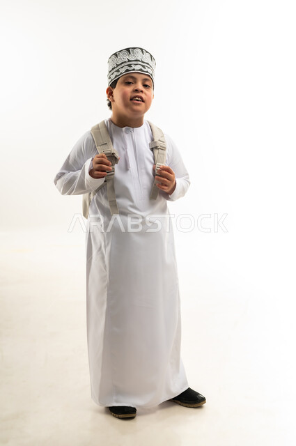 دمج ذوي الاحتياجات الخاصة في المدارس، الإستعداد للعام الدراسي الجديد، بورتريه لولد عربي خليجي عماني مبتسم مصاب بمتلازمة داون يرتدي الدشداشة والكمة يحمل حقيبة الظهر ينظر الى الكاميرا بايماءات السرور، صورة لكامل الجسم، خلفية بيضاء