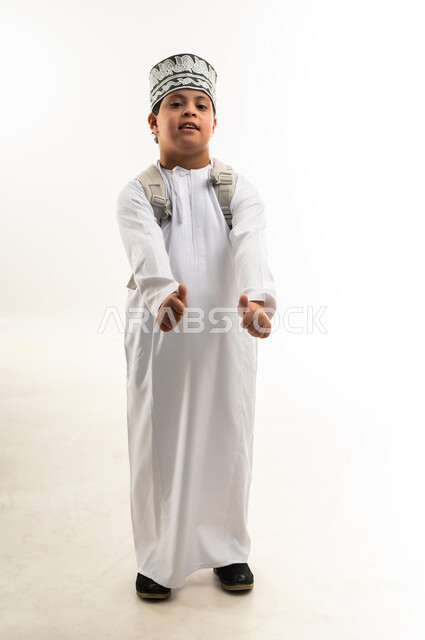 تعابير القبول والموافقة، دمج ذوي الاحتياجات الخاصة في المدارس، الإستعداد للعام الدراسي الجديد، بورتريه لولد عربي خليجي عماني مبتسم مصاب بمتلازمة داون يرتدي الدشداشة والكمة يحمل حقيبة الظهر يرفع يديه بايماءات الاعجاب والرضى، صورة لكامل الجسم، خلفية بيضاء