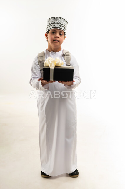 التعبير عن الألفة والمحبة، تقديم الهدايا في الأعياد والمناسبات، دمج ذوي الاحتياجات الخاصة في المجتمع العماني، بورتريه لصبي عربي خليجي عماني يرتدي الدشداشة والكمة و حقيبة الظهر المدرسية يحمل علبة هدية ملونة، صورة لكامل الجسم، خلفية بيضاء