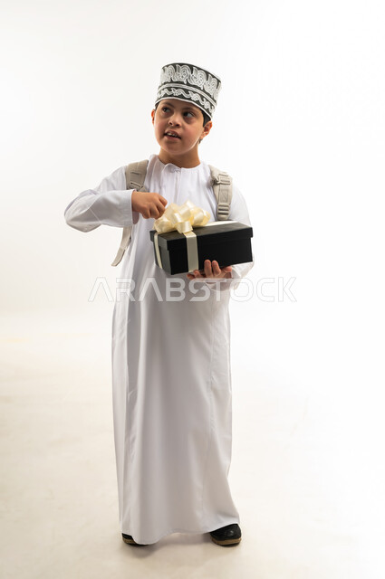 تقديم الهدايا في الأعياد والمناسبات، التعبير عن الألفة والمحبة، دمج ذوي الاحتياجات الخاصة في المجتمع العماني، بورتريه لصبي عربي خليجي عماني يرتدي الدشداشة والكمة و حقيبة الظهر المدرسية يحمل علبة هدية ملونة، صورة لكامل الجسم، خلفية بيضاء