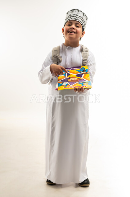 النظر الى الكاميرا بايماءات السرور، الإستعداد للعام الدراسي الجديد، بورتريه لولد عربي خليجي عماني مبتسم مصاب بمتلازمة داون يرتدي الدشداشة والكمة يحمل الحقيبة والكتب المدرسية يشير باصبعه لكتابه، دمج ذوي الاحتياجات الخاصة في المدارس، صورة لكامل الجسم، خلفية بيضاء