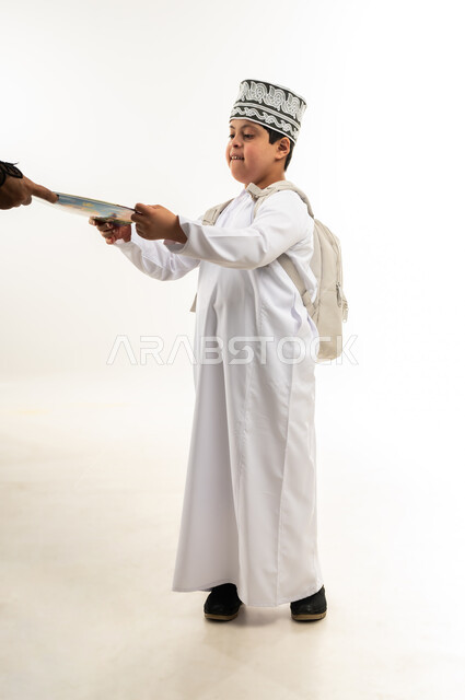 دمج ذوي الاحتياجات الخاصة في المدارس، الإستعداد للعام الدراسي الجديد، بورتريه من الجانب لولد عربي خليجي عماني مبتسم مصاب بمتلازمة داون يرتدي الدشداشة والكمة يحمل الحقيبة يقوم باستلام الكتب المدرسية من معلمته، صورة لكامل الجسم، خلفية بيضاء