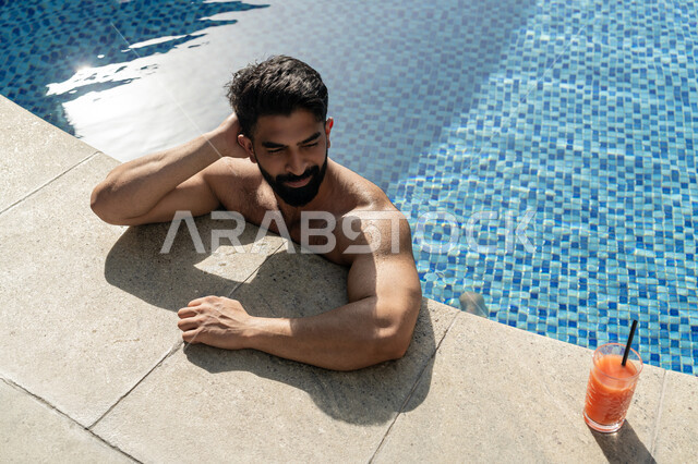 القيام بالأنشطة الترفيهية المسلية, مفهوم الاستجمام والاختلاء, الاستمتاع بالمشروبات البارد المنعشة, ممارسة رياضة السباحة الممتعة, الجلوس والاسترخاء في المسبح, شاب عربي خليجي إماراتي يجلس داخل حوض السباحة في إحدى المنتجعات السياحية بالإمارات