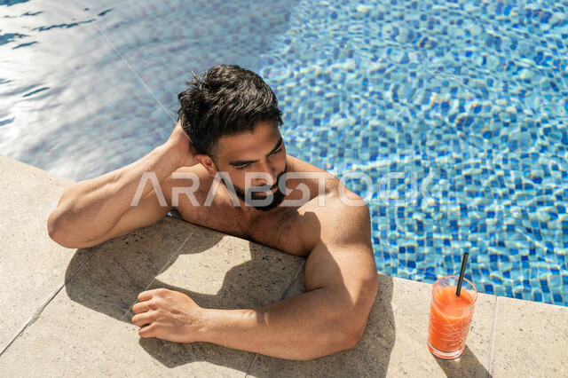 الاستمتاع بالمشروبات البارد المنعشة, ممارسة رياضة السباحة الممتعة, الجلوس والاسترخاء في المسبح, مفهوم الاستجمام والاختلاء, شاب عربي خليجي إماراتي يجلس داخل حوض السباحة في إحدى المنتجعات السياحية بالإمارات, القيام بالأنشطة الترفيهية المسلية
