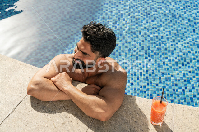 القيام بالأنشطة الترفيهية المسلية, مفهوم الاستجمام والاختلاء, الاستمتاع بالمشروبات البارد المنعشة, ممارسة رياضة السباحة الممتعة, الجلوس والاسترخاء في المسبح, شاب عربي خليجي إماراتي يجلس داخل حوض السباحة في إحدى المنتجعات السياحية بالإمارات