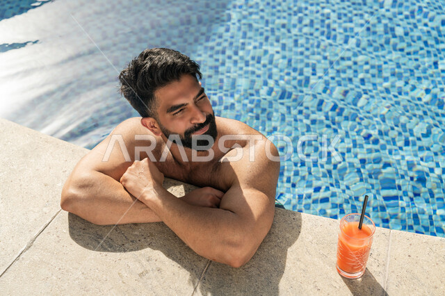 الجلوس والاسترخاء في المسبح, ممارسة رياضة السباحة الممتعة, القيام بالأنشطة الترفيهية المسلية, مفهوم الاستجمام والاختلاء, شاب عربي خليجي إماراتي يجلس داخل حوض السباحة في إحدى المنتجعات السياحية بالإمارات, الاستمتاع بالمشروبات البارد المنعشة