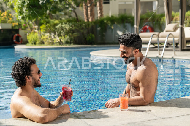 قضاء العطلة الصيفية في المنتجعات السياحية, مشاركة الأصدقاء اللحظات الممتعة, القيام بالأنشطة الترفيهية المسلية, شابان عربيان خليجيان إماراتيان يجلسان داخل حوض السباحة في إحدى الفنادق بالإمارات, الاستمتاع بالمشروبات الباردة المنعشة