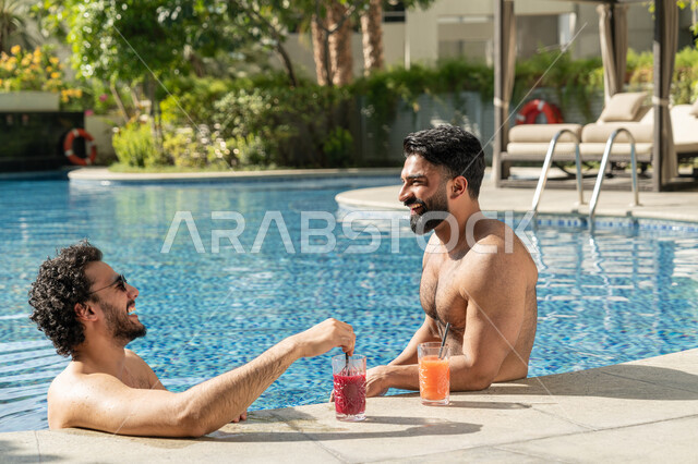 القيام بالأنشطة الترفيهية المسلية, مشاركة الأصدقاء اللحظات الممتعة, قضاء العطلة الصيفية في المنتجعات السياحية, شابان عربيان خليجيان إماراتيان يجلسان داخل حوض السباحة في إحدى الفنادق بالإمارات, الاستمتاع بالمشروبات الباردة المنعشة