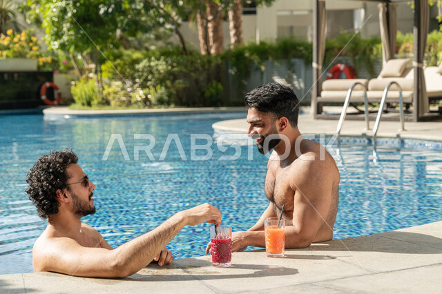 قضاء العطلة الصيفية في المنتجعات السياحية, مشاركة الأصدقاء اللحظات الممتعة, القيام بالأنشطة الترفيهية المسلية, شابان عربيان خليجيان إماراتيان يجلسان داخل حوض السباحة في إحدى الفنادق بالإمارات, الاستمتاع بالمشروبات الباردة المنعشة