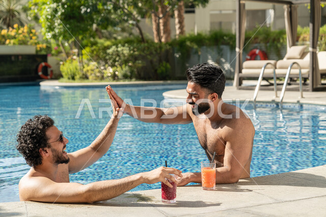 مشاركة الأصدقاء اللحظات الممتعة, قضاء العطلة الصيفية في المنتجعات السياحية, القيام بالأنشطة الترفيهية المسلية, شابان عربيان خليجيان إماراتيان يجلسان داخل حوض السباحة في إحدى الفنادق بالإمارات, الاستمتاع بالمشروبات الباردة المنعشة