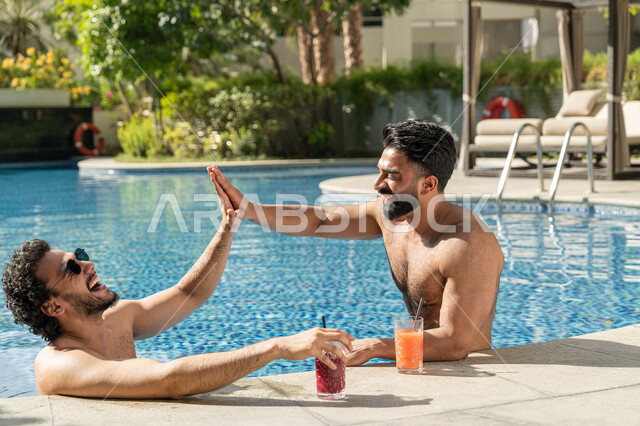 القيام بالأنشطة الترفيهية المسلية, مشاركة الأصدقاء اللحظات الممتعة, قضاء العطلة الصيفية في المنتجعات السياحية, شابان عربيان خليجيان إماراتيان يجلسان داخل حوض السباحة في إحدى الفنادق بالإمارات, الاستمتاع بالمشروبات الباردة المنعشة